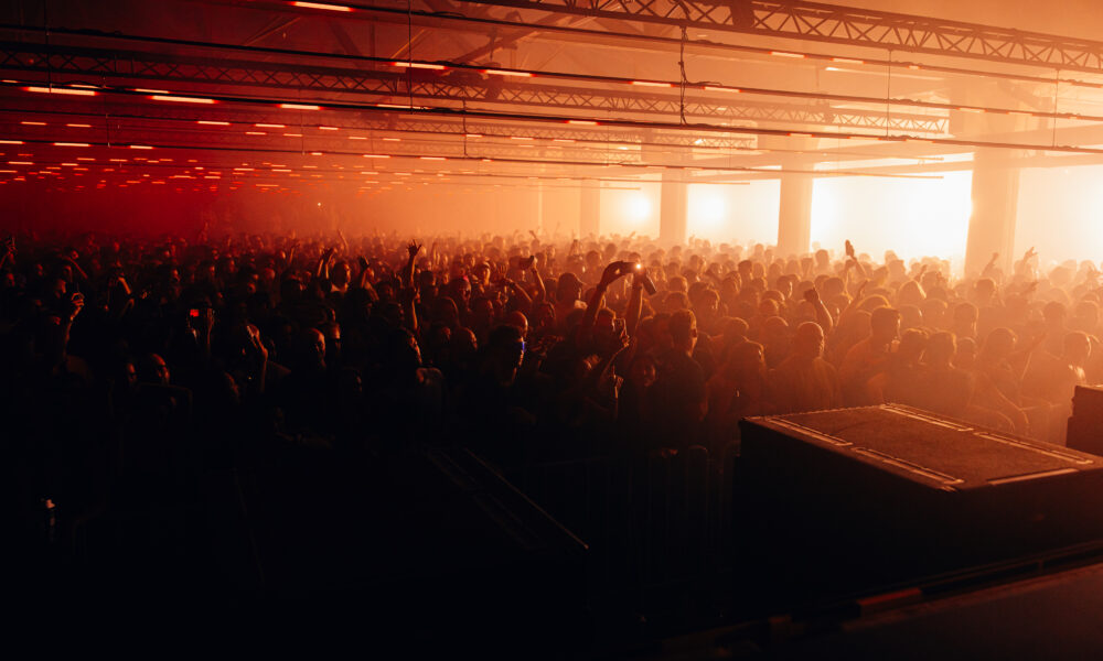 A high-energy night shot of a massive crowd at Audio Obscura Festival in Rotterdam, with industrial buildings and brutalist architecture illuminated by pink and purple stage lights.