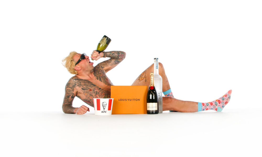 CASH ONLY, heavily tattooed and shirtless, reclines on a white studio floor drinking from a wine bottle, surrounded by a KFC bucket, a Louis Vuitton box, Moët & Chandon champagne, and a Grey Goose bottle, wearing pink patterned socks and dark sunglasses.