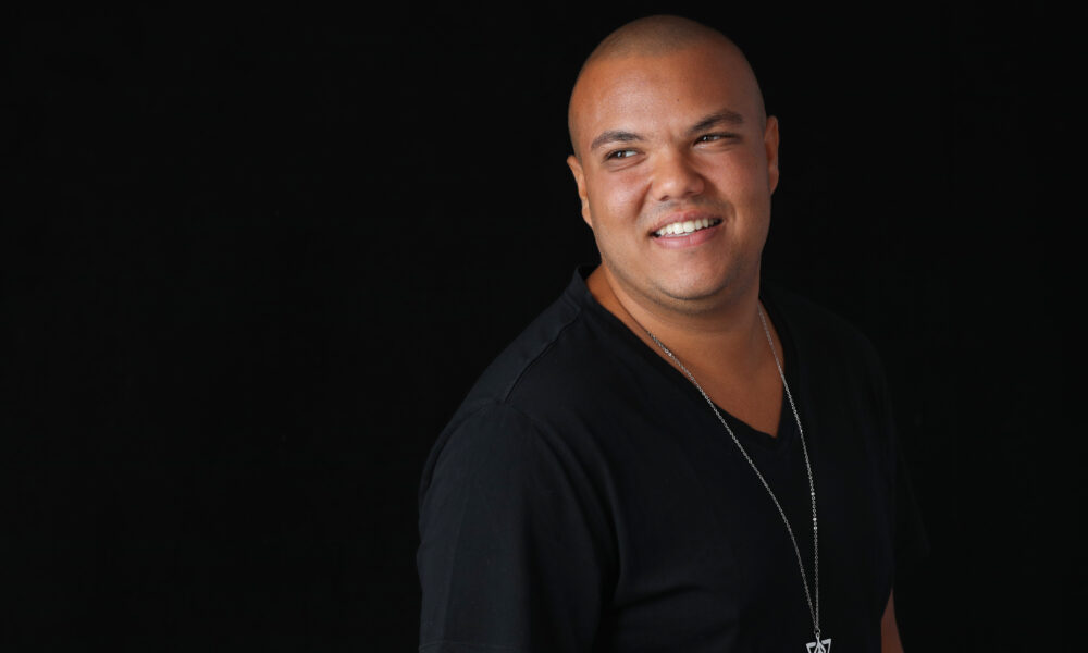 ALTRIGO, the Egyptian-born Los Angeles-based DJ and producer, smiling over his shoulder in a studio portrait wearing a black v-neck t-shirt and silver geometric pendant necklace against a dark black background.