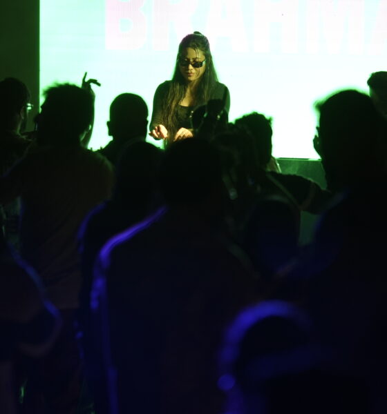 Brahma (IN) performing behind the decks at a darkened club night, wearing dark sunglasses and a black outfit, lit from above by green stage lighting with a bright white projection screen visible behind her and a crowd of silhouetted heads in the foreground.