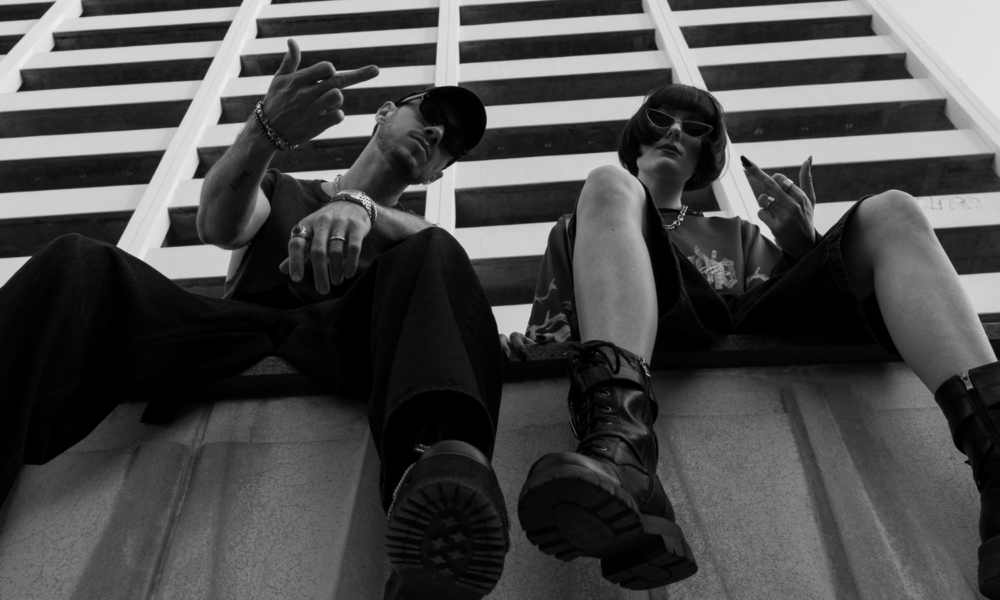 Black-and-white low-angle promotional photo of an electronic music duo wearing sunglasses and dark streetwear while seated beneath a geometric building facade.