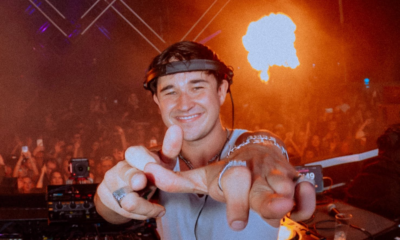 John Summit smiling and pointing at the camera while DJing in front of a large crowd bathed in orange light