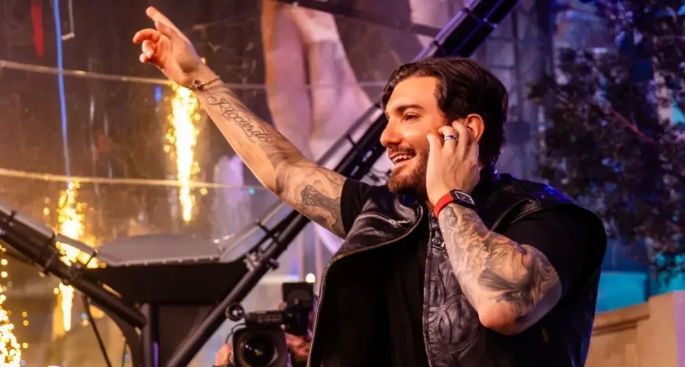 Alesso smiling and pointing to the crowd while DJing on stage with warm stage lighting behind him
