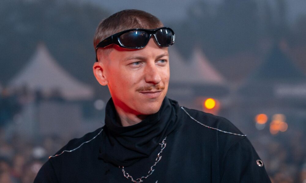 Björn Grimm, co-founder of Nibirii Festival, standing at the DJ booth wearing sunglasses and a black turtleneck top with festival crowds in the background