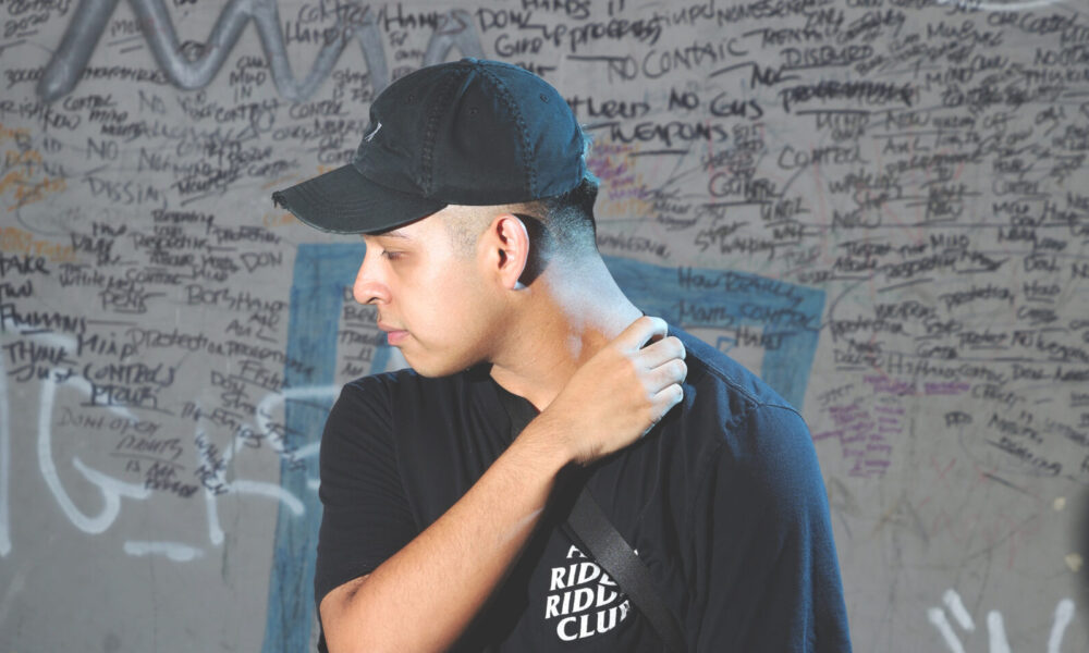 Deadlynoiz wearing a black cap and t-shirt reading "Ridin Ridin Club" standing against a graffiti-covered wall