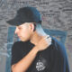 Deadlynoiz wearing a black cap and t-shirt reading "Ridin Ridin Club" standing against a graffiti-covered wall