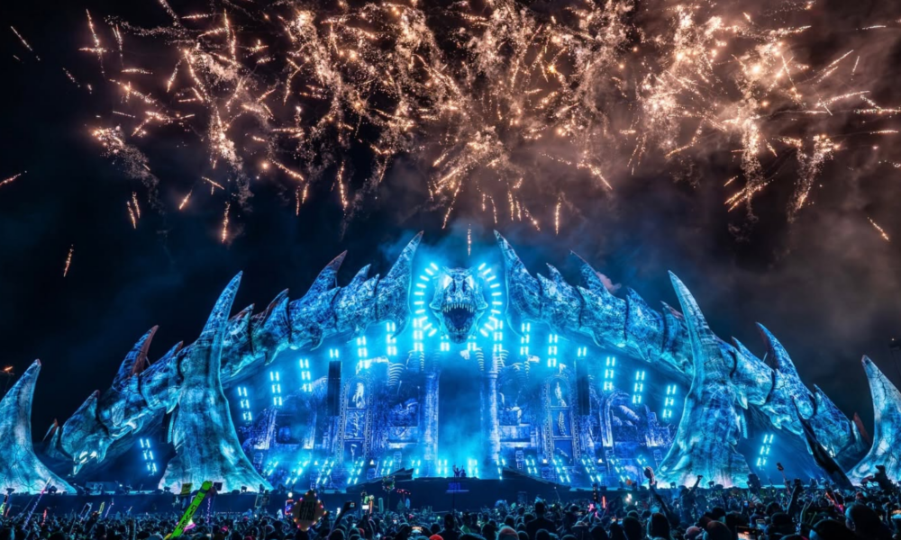 Lost Lands Festival at night with its large dinosaur-themed stage lit in blue as fireworks fill the sky above the crowd.