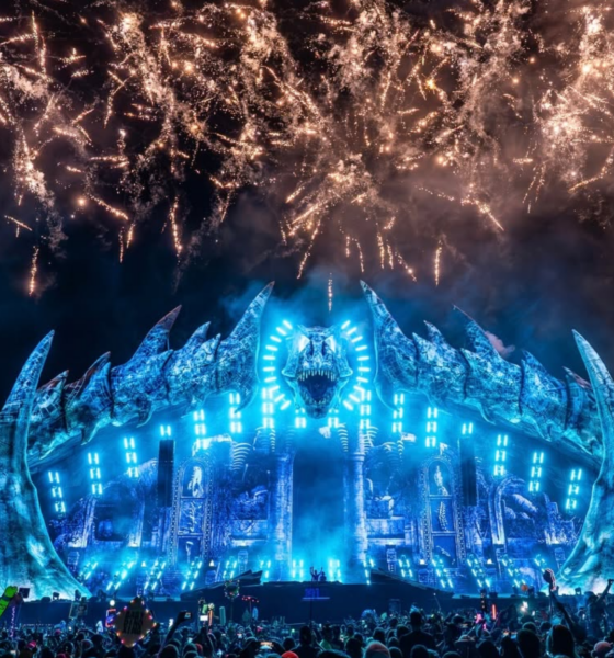 Lost Lands Festival at night with its large dinosaur-themed stage lit in blue as fireworks fill the sky above the crowd.