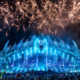 Lost Lands Festival at night with its large dinosaur-themed stage lit in blue as fireworks fill the sky above the crowd.
