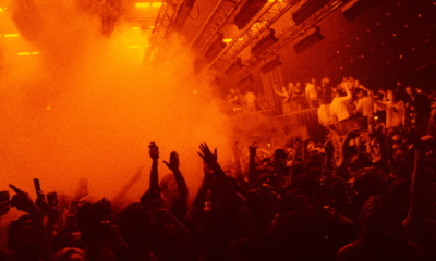A packed crowd inside Hï Ibiza during a live DJ set, with orange lighting and smoke filling the club as people dance around the booth.
