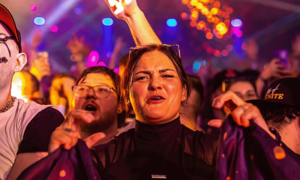 A woman at an electronic music event pulling a scrunched bass face while reacting to the music, surrounded by other people under colourful stage lighting.
