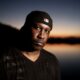 Todd Terry wearing a black beanie, standing against a sunset sky over water