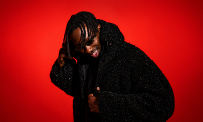 AMÉMÉ smiling and adjusting his sunglasses against a vivid red backdrop, wearing a black textured jacket