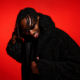 AMÉMÉ smiling and adjusting his sunglasses against a vivid red backdrop, wearing a black textured jacket