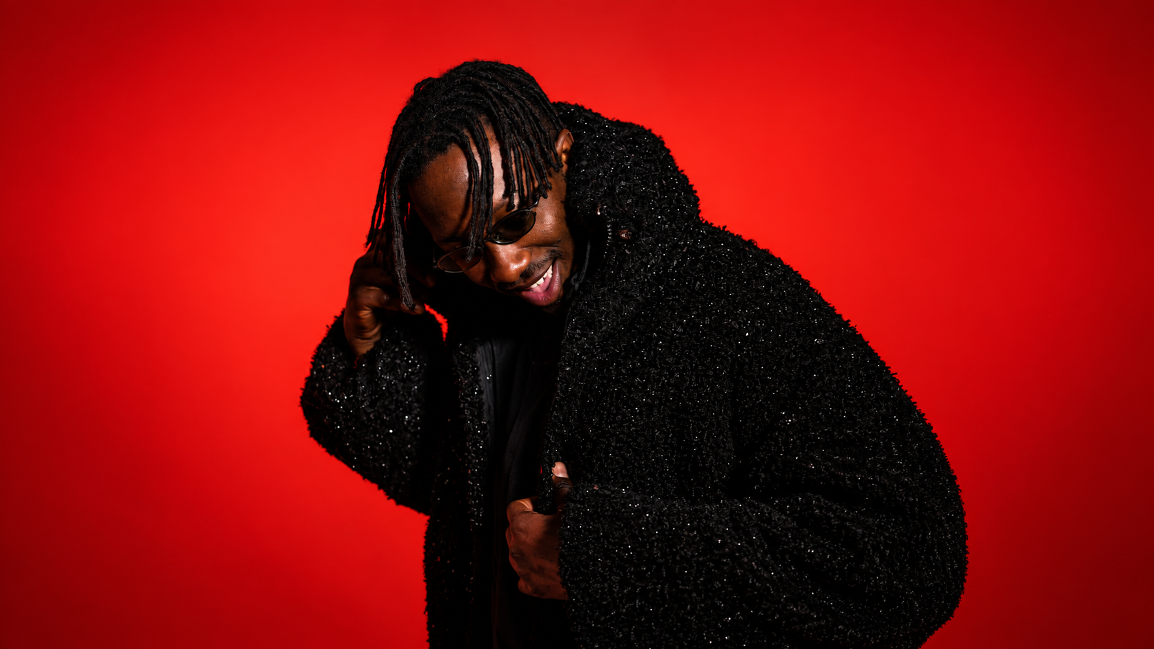 AMÉMÉ smiling and adjusting his sunglasses against a vivid red backdrop, wearing a black textured jacket