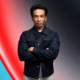 Laidback Luke wearing a denim jacket with arms crossed against a red and teal abstract background