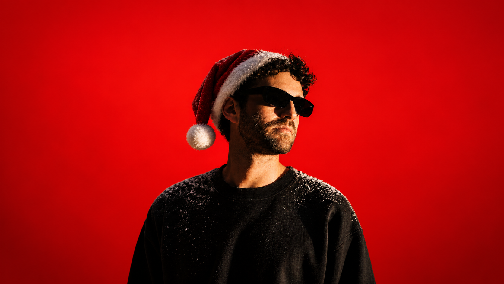 DJ wearing sunglasses and a Santa hat against a bright red background, dusted with snow on his shoulders