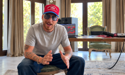 HUGEL sitting in a home studio wearing a red cap and sunglasses, gesturing while speaking to camera, with production equipment visible behind him