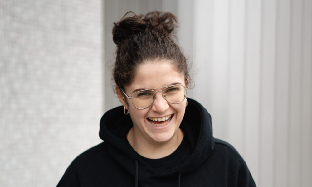 A.N.I. laughing while wearing a black hoodie with a red ace of hearts embroidery, standing against a striped grey backdrop