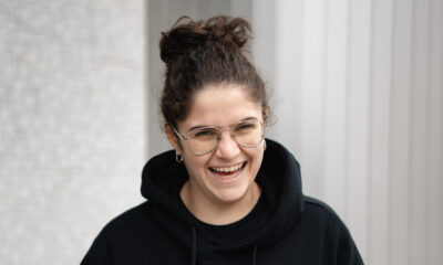 A.N.I. laughing while wearing a black hoodie with a red ace of hearts embroidery, standing against a striped grey backdrop
