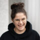 A.N.I. laughing while wearing a black hoodie with a red ace of hearts embroidery, standing against a striped grey backdrop