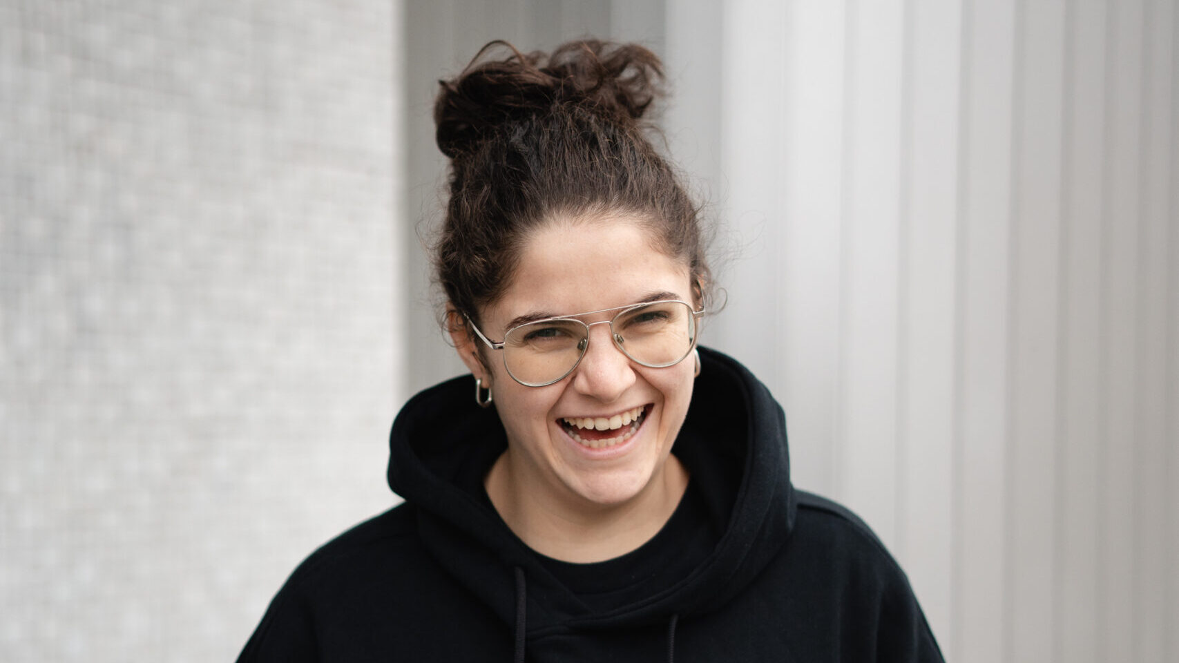 A.N.I. laughing while wearing a black hoodie with a red ace of hearts embroidery, standing against a striped grey backdrop