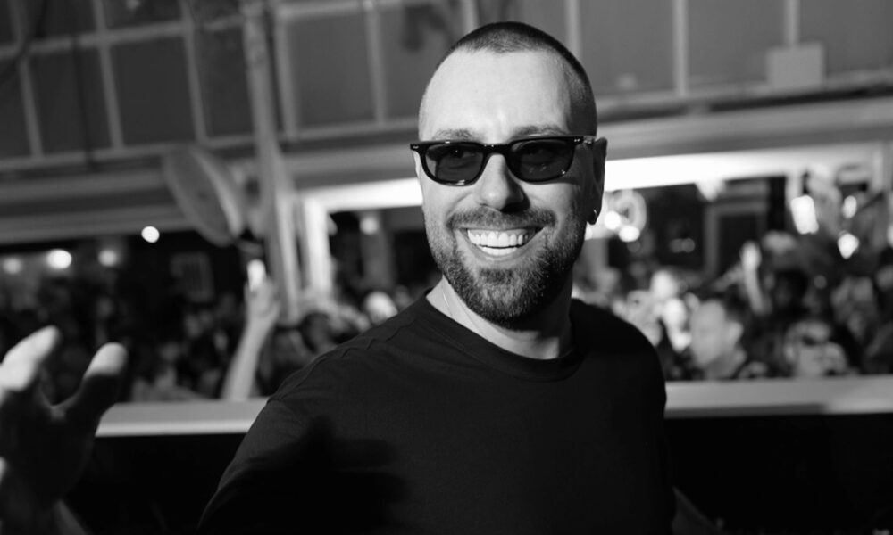 Black and white photo of Sebastian Ingrosso smiling and reaching toward the camera in front of a crowd
