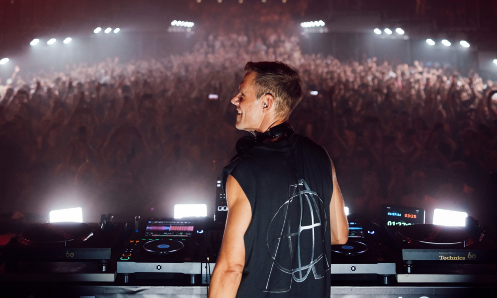 Armin van Buuren performing behind the decks in front of a large crowd during a live show at UNVRS