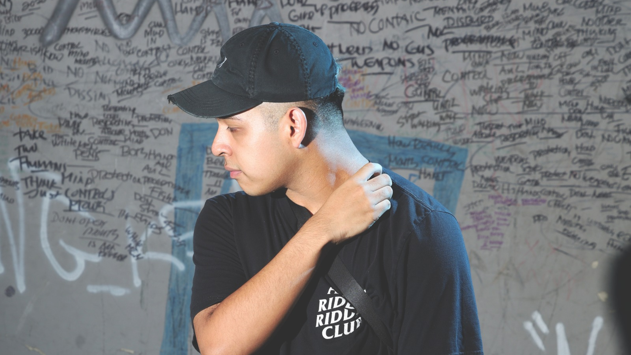Deadlynoiz wearing a black cap and black shirt in front of a graffiti-covered wall