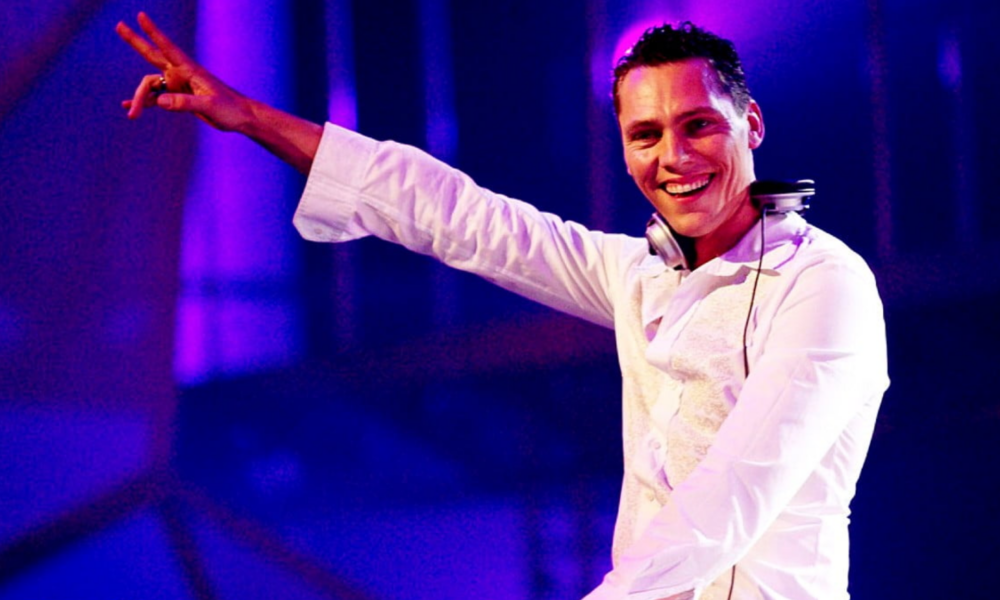 Tiësto performing on stage in a white outfit with headphones around his neck and his arm raised under blue and purple lighting