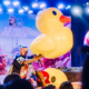 Lenny Pearce performing at a baby rave with a giant inflatable duck, colorful stage decorations and families watching from the crowd