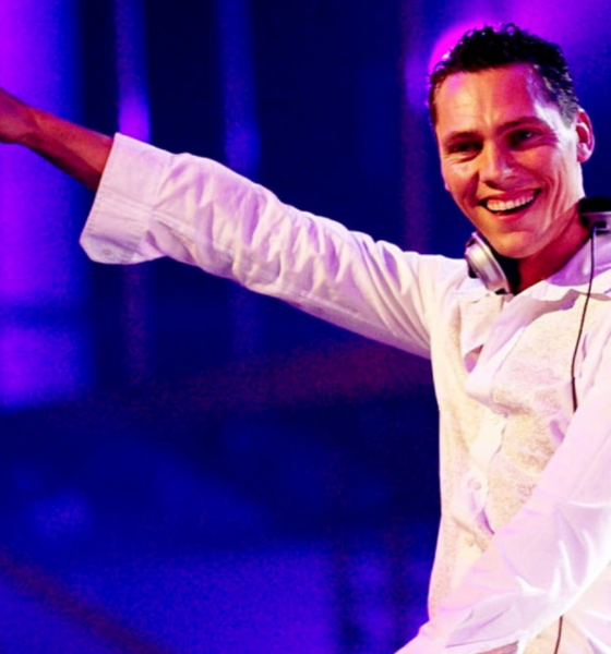 Tiësto performing on stage in a white outfit with headphones around his neck and his arm raised under blue and purple lighting