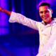 Tiësto performing on stage in a white outfit with headphones around his neck and his arm raised under blue and purple lighting