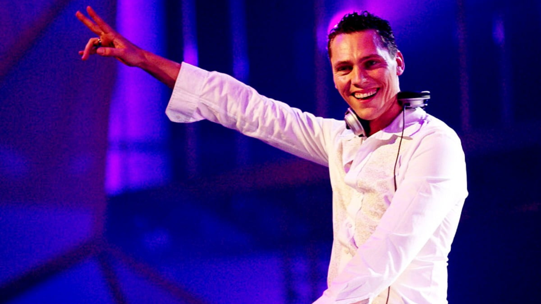 Tiësto performing on stage in a white outfit with headphones around his neck and his arm raised under blue and purple lighting