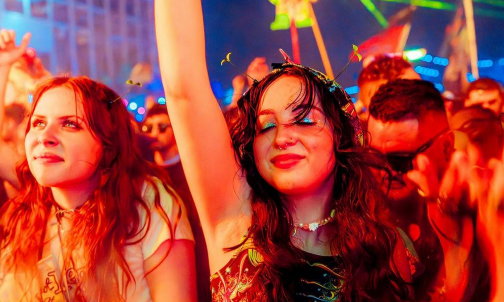Close-up of two women with eyes closed and arms raised, dancing joyfully in a crowd bathed in red festival lighting, expressing emotional release on the dancefloor.