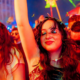 Close-up of two women with eyes closed and arms raised, dancing joyfully in a crowd bathed in red festival lighting, expressing emotional release on the dancefloor.