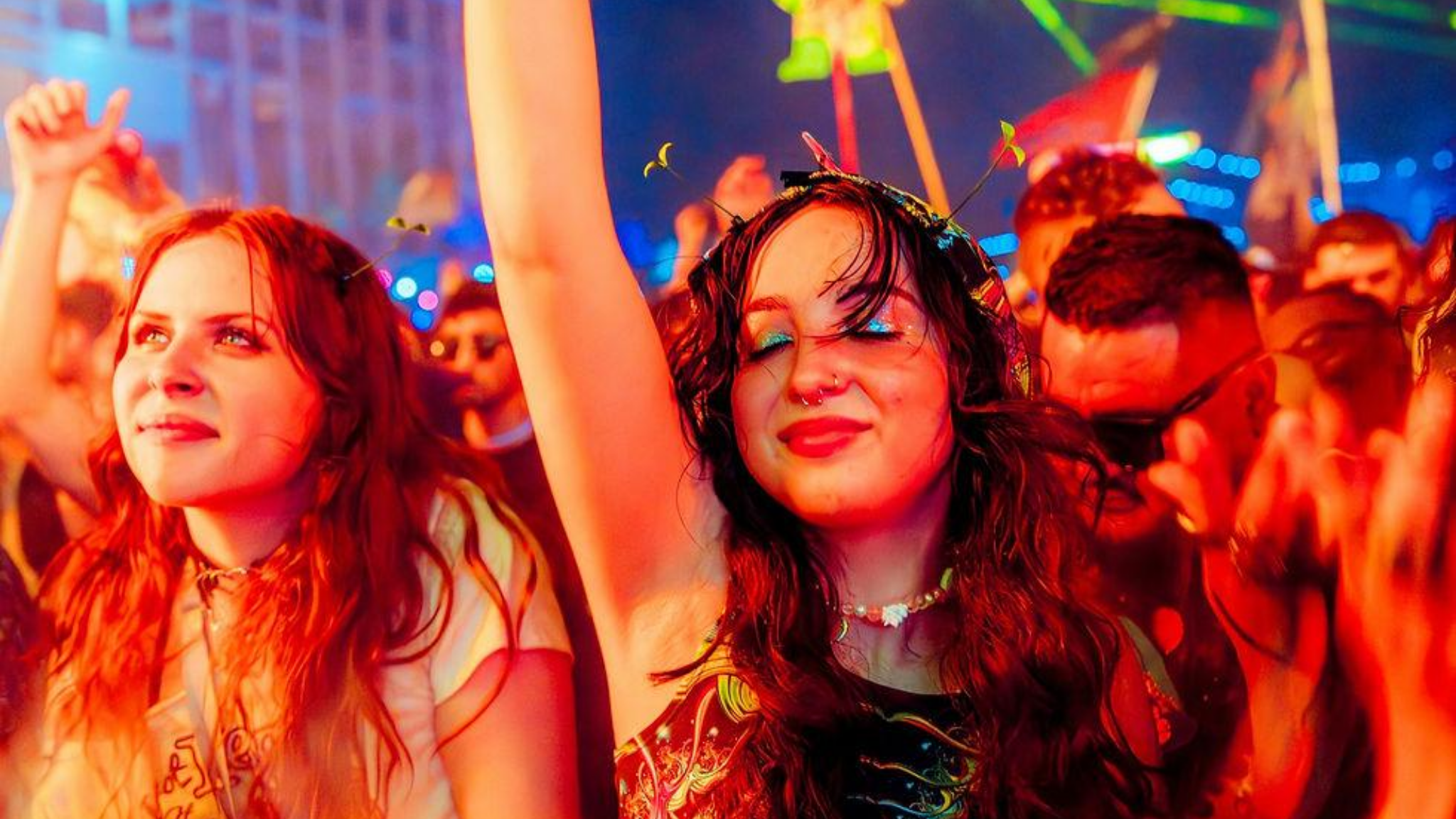 Close-up of two women with eyes closed and arms raised, dancing joyfully in a crowd bathed in red festival lighting, expressing emotional release on the dancefloor.