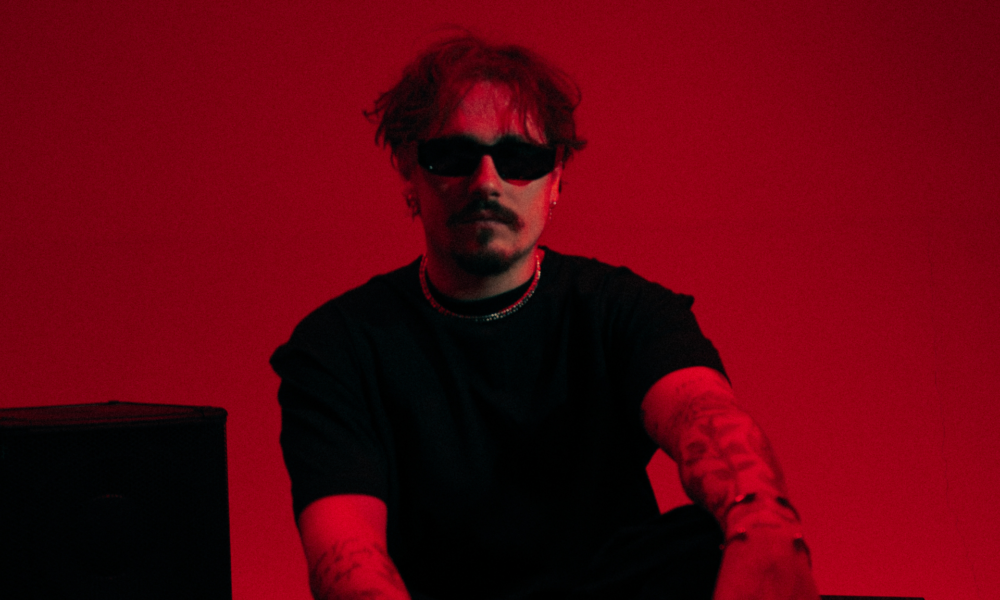 REYUS wearing black sunglasses and a black shirt against a red background ahead of the release of ‘Deeper Water’ with Émilie Rachel.