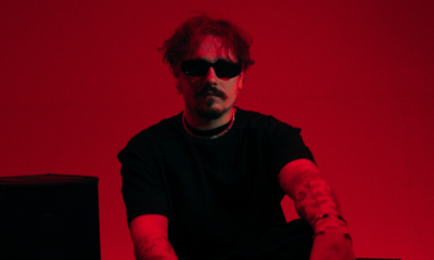 REYUS wearing black sunglasses and a black shirt against a red background ahead of the release of ‘Deeper Water’ with Émilie Rachel.