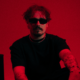 REYUS wearing black sunglasses and a black shirt against a red background ahead of the release of ‘Deeper Water’ with Émilie Rachel.