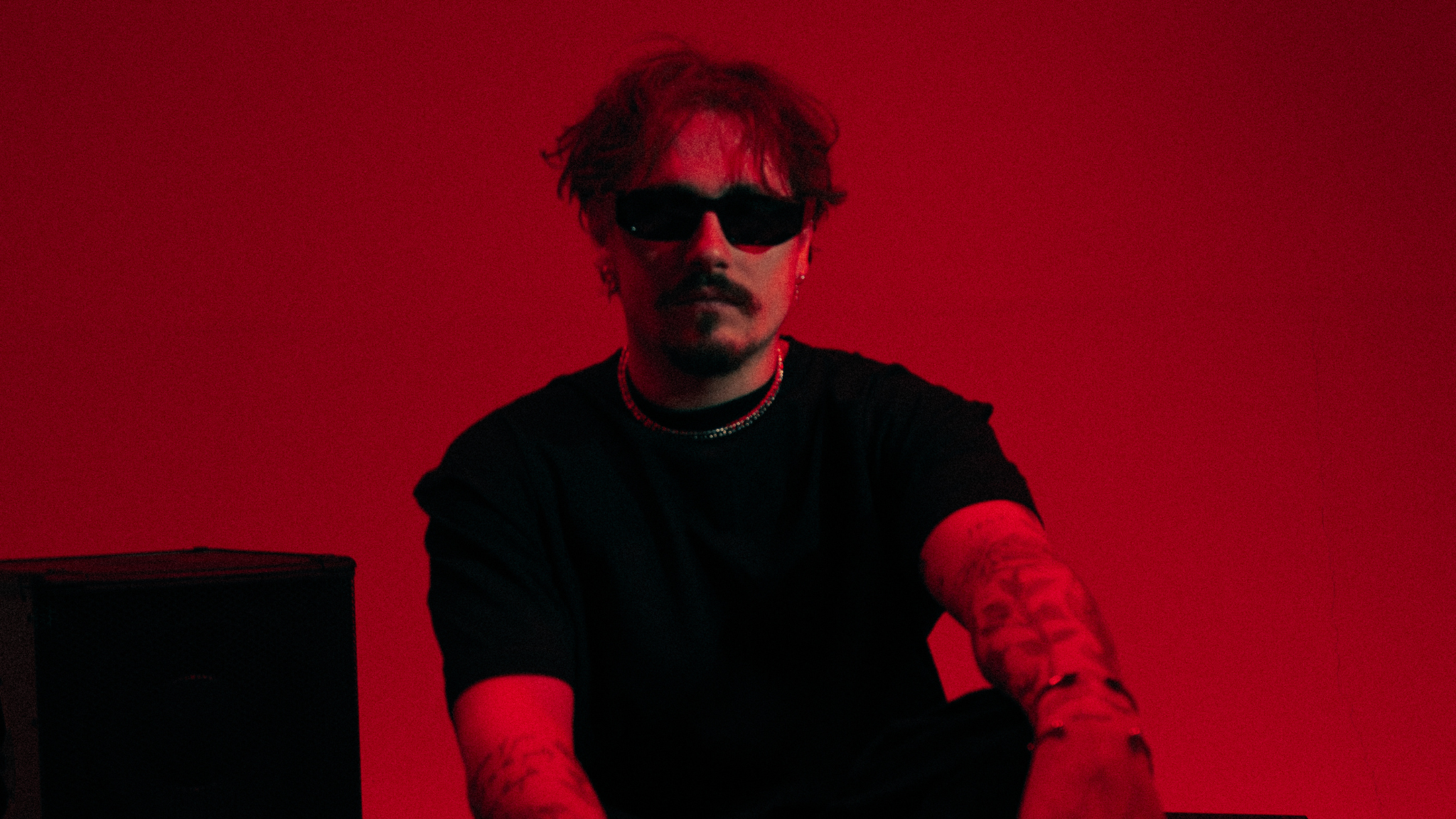 REYUS wearing black sunglasses and a black shirt against a red background ahead of the release of ‘Deeper Water’ with Émilie Rachel.