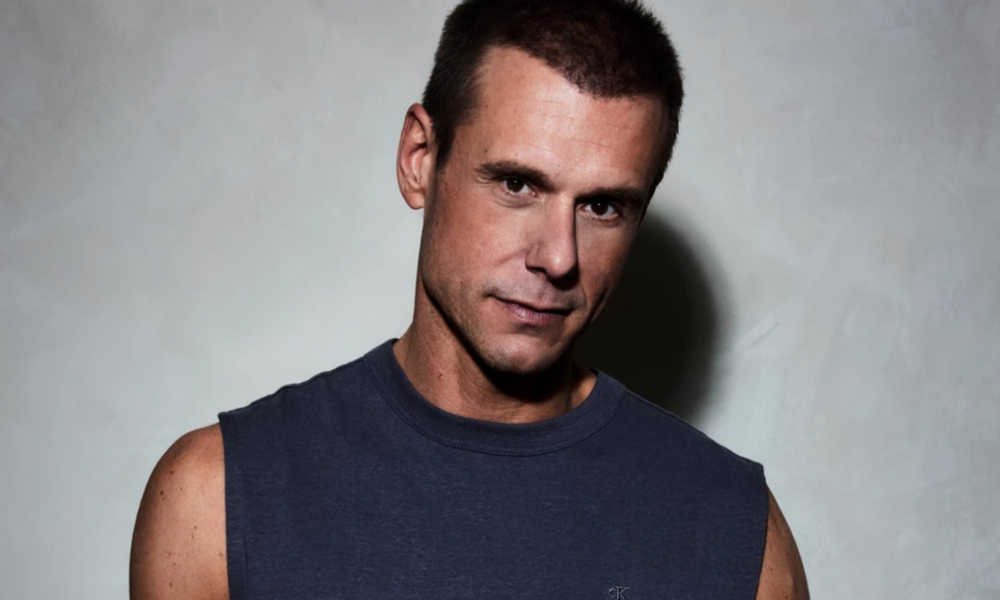 Armin van Buuren wearing a sleeveless dark shirt while posing against a light grey background