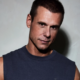 Armin van Buuren wearing a sleeveless dark shirt while posing against a light grey background