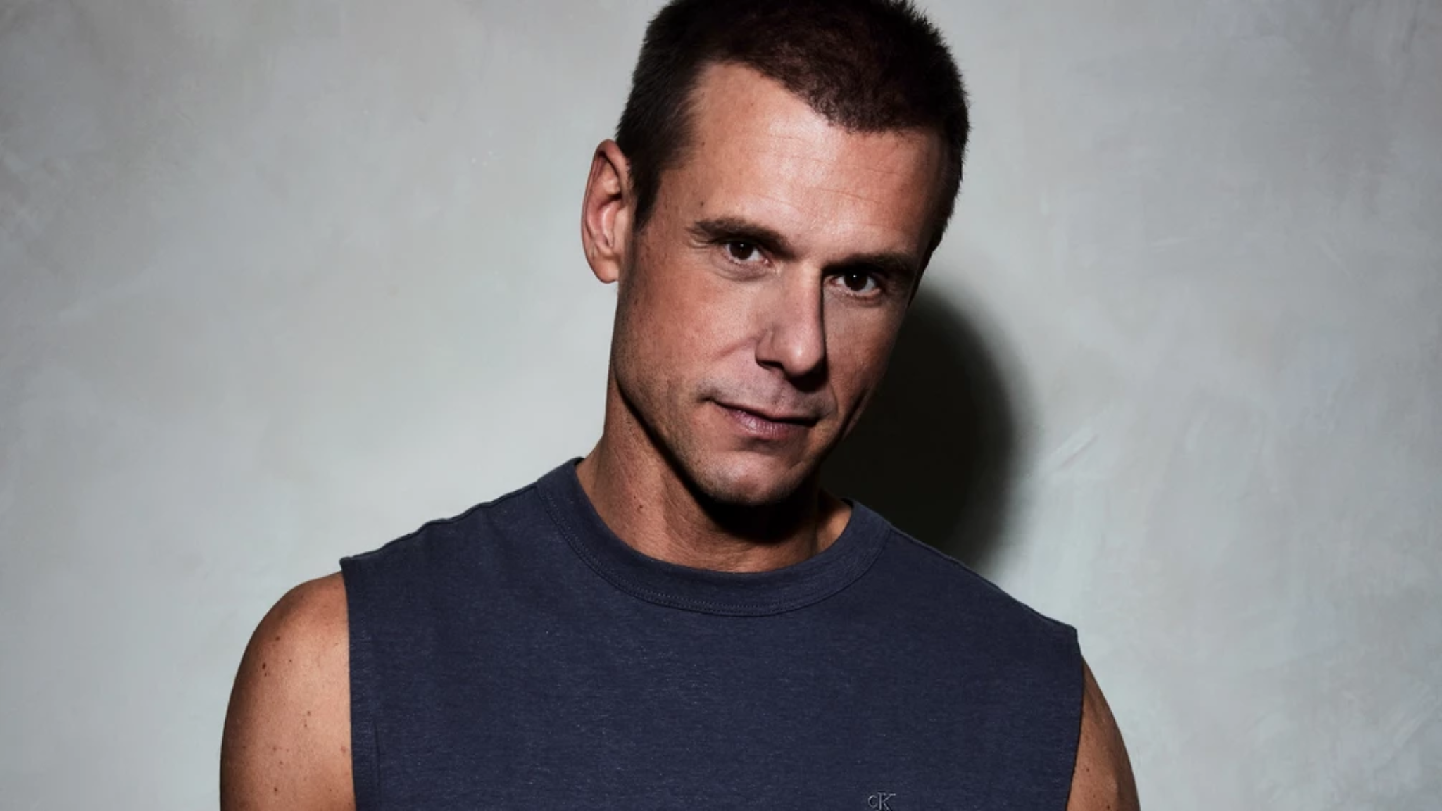 Armin van Buuren wearing a sleeveless dark shirt while posing against a light grey background