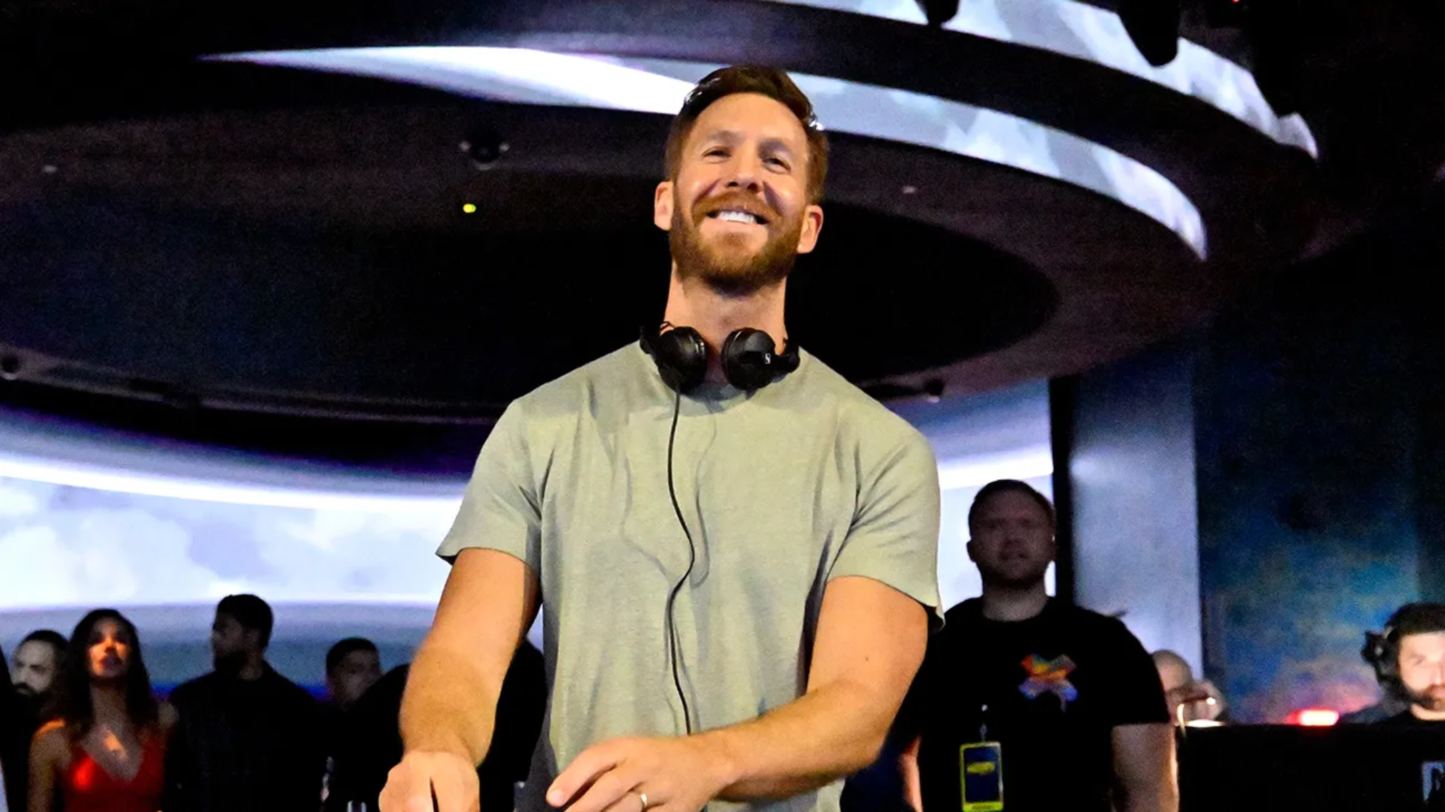 Calvin Harris smiling behind the decks with headphones around his neck during a live performance