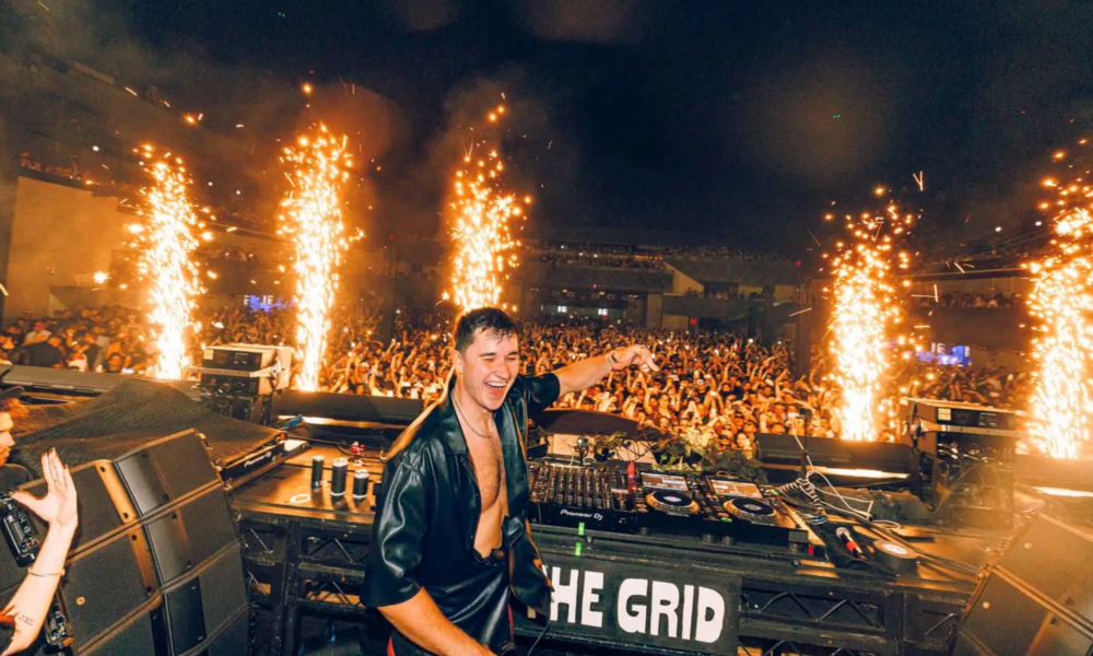 John Summit performing behind the decks in front of a large crowd as pyrotechnics fire around the stage
