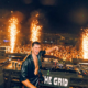 John Summit performing behind the decks in front of a large crowd as pyrotechnics fire around the stage