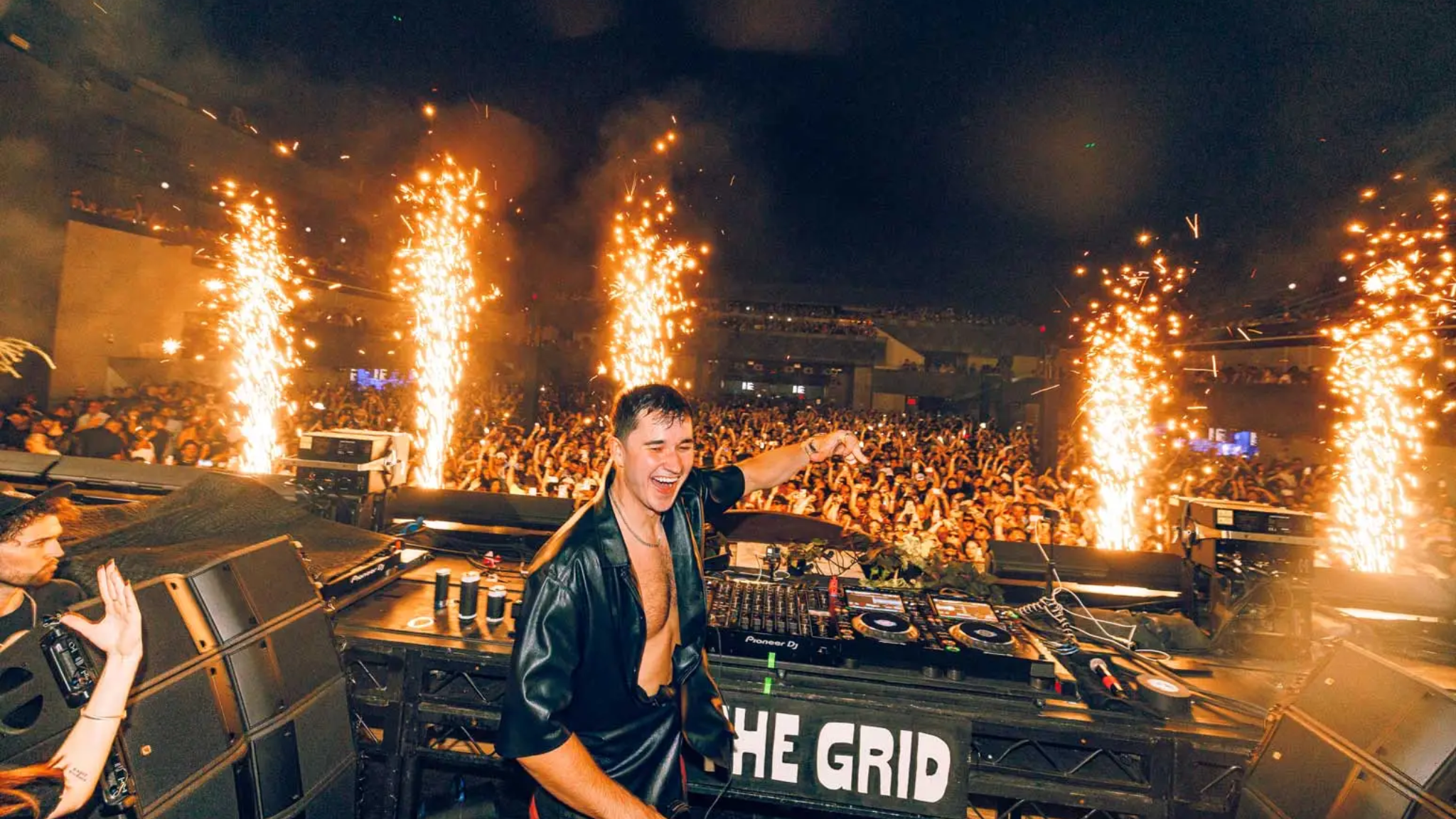 John Summit performing behind the decks in front of a large crowd as pyrotechnics fire around the stage