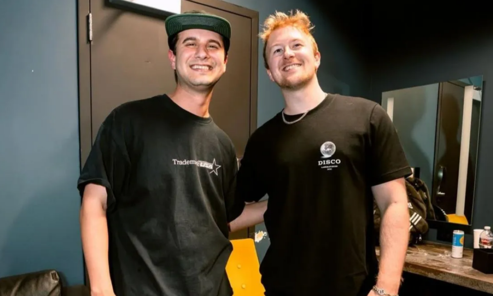 Tape B and Effin smiling together in a dressing room, both wearing black T-shirts, with mirrors and a counter visible behind them