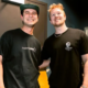 Tape B and Effin smiling together in a dressing room, both wearing black T-shirts, with mirrors and a counter visible behind them